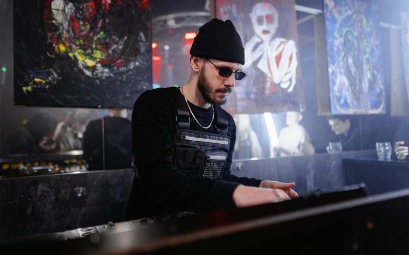 Energetic DJ in sunglasses spins tracks at a lively indoor club event with abstract art.